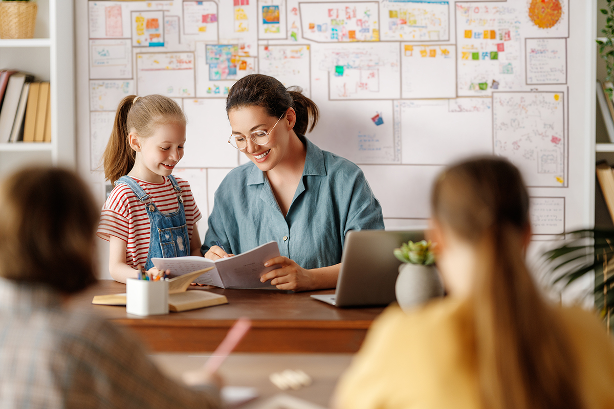 Teacher sitting at her desk resolves the doubts of the girl standing next to her. 