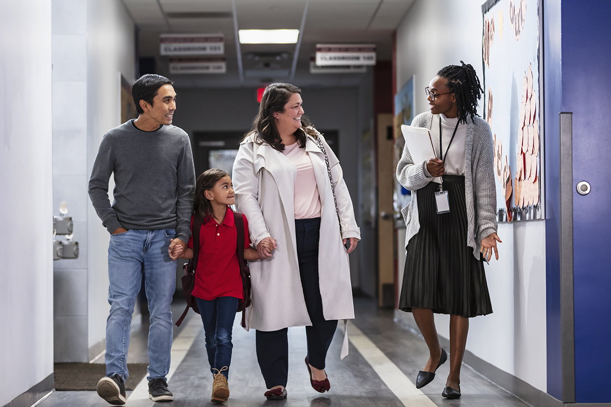 Two parents take their daughter to school and talk to a primary school teacher in the hallway.