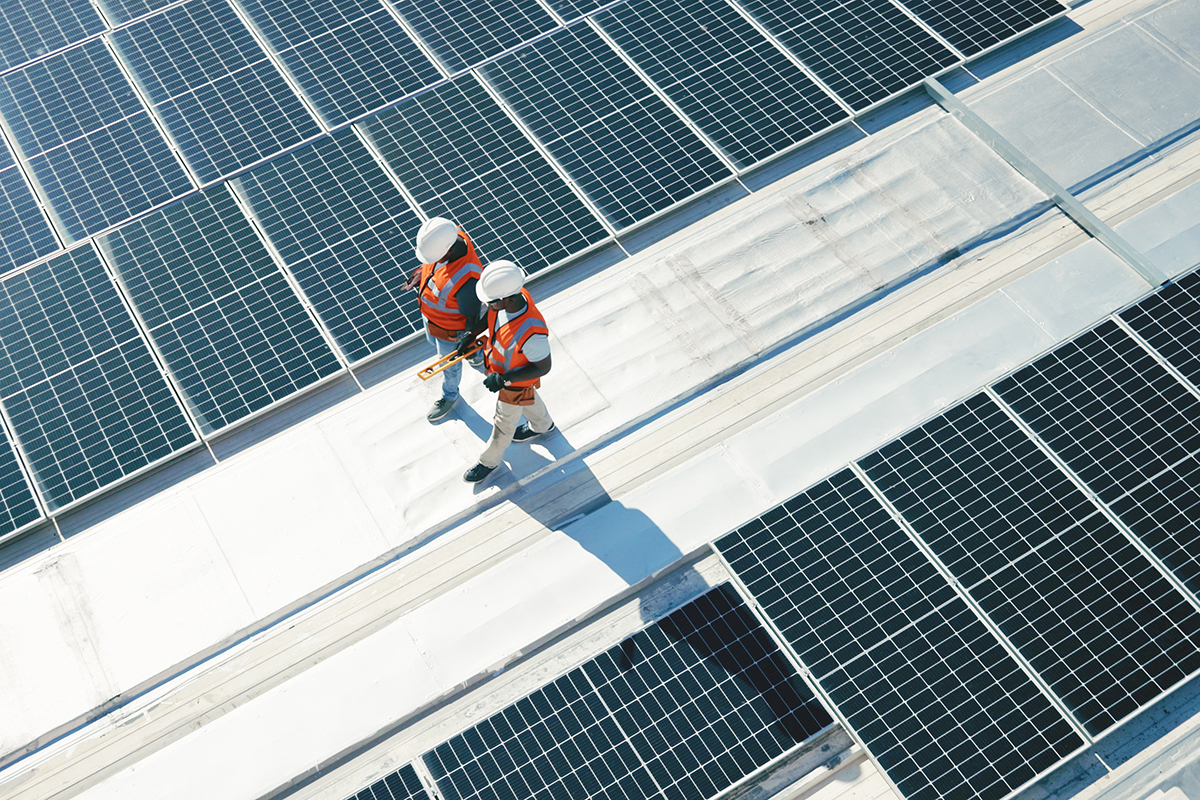 Trabalhadores da construção civil caminham em meio a painéis solares instalados no telhado de um edifício. 