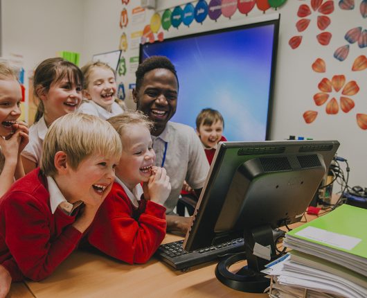 Um grupo de alunos do ensino fundamental está reunido em torno de um computador com seu professor. Todos estão rindo e olhando para a tela do computador.