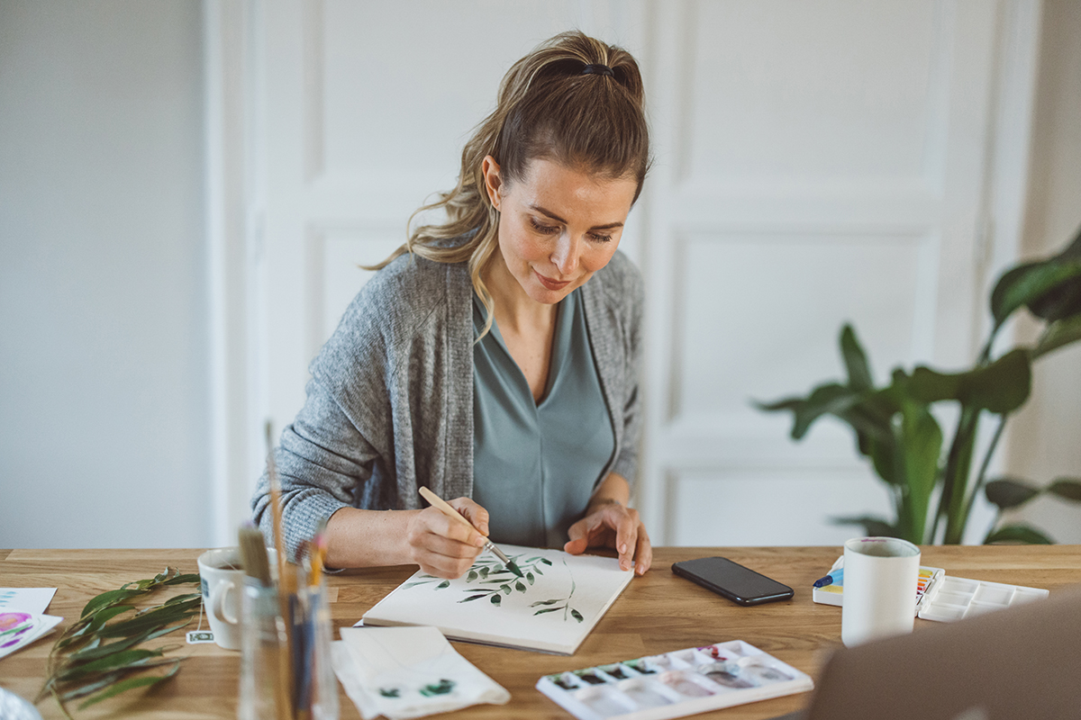 Mulher madura em casa pintando. Ela está assistindo a um tutorial no computador e pintando. Sentada no balcão da cozinha em frente à janela.