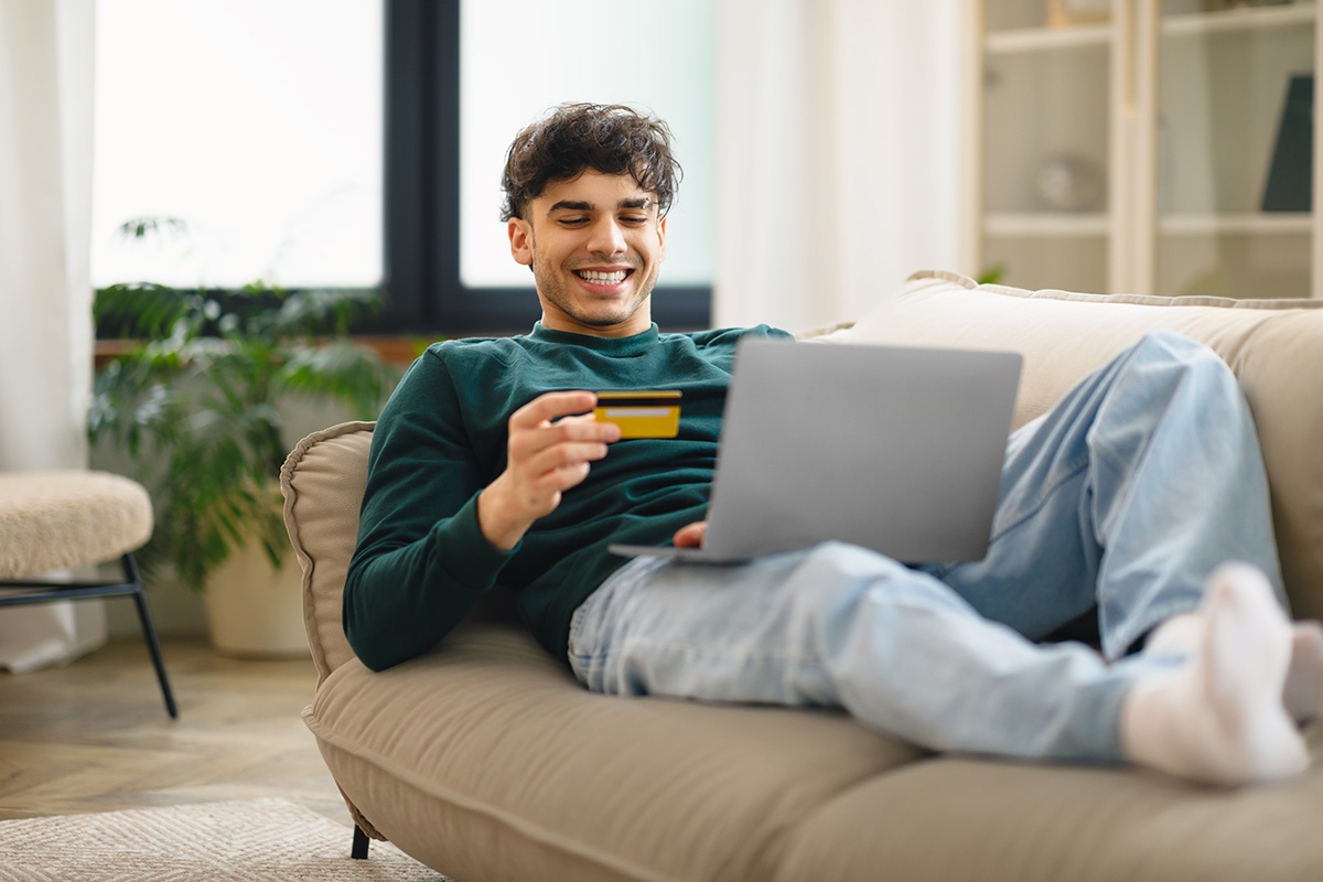 Homem jovem fazendo compras on-line. Recostado no sofá, ele usa seu laptop e segura seu cartão de crédito. 