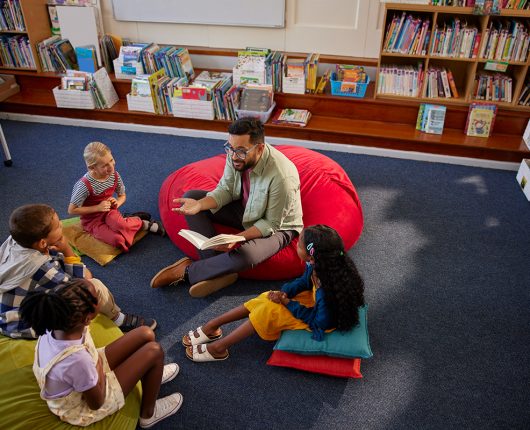 Alunos curiosos do ensino fundamental sentam-se em uma grande almofada na biblioteca enquanto ouvem uma história do bibliotecário. 