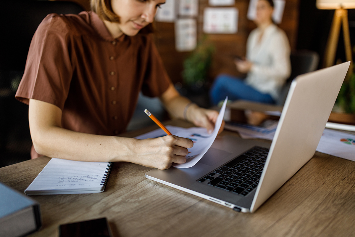 Retrato de uma jovem diligente analisando documentos, fazendo anotações e usando um laptop em sua mesa em um escritório moderno.