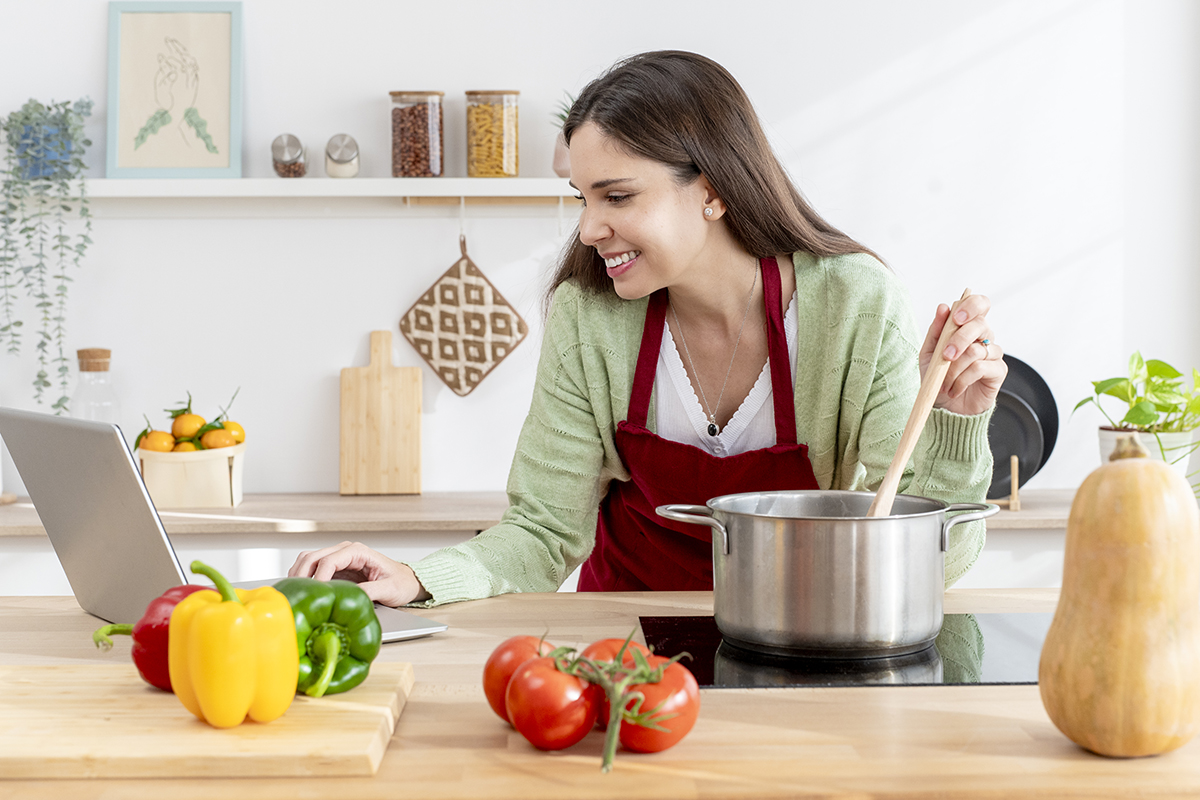 Dona de casa feliz mexendo uma panela e lendo uma receita em um laptop cercada por vegetais frescos em uma cozinha moderna