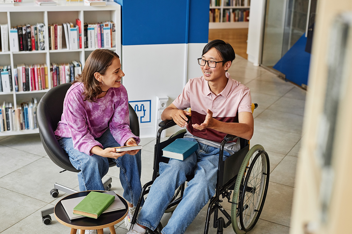 Joven estudiante  en silla de ruedas charla con otra compañera en la biblioteca. 