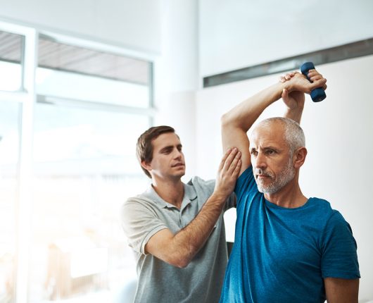 Adulto mayor realiza ejercicios con peso mientras es supervisado por un experto deportivo. 