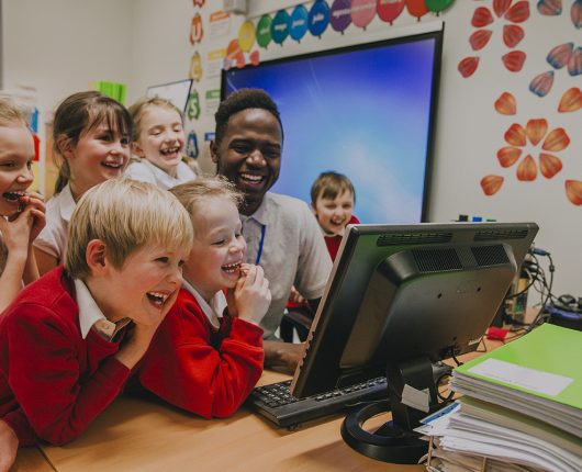 Un grupo de alumnos de primaria se apiñan alrededor de un ordenador con su profesor. Todos ríen y miran la pantalla del ordenador.