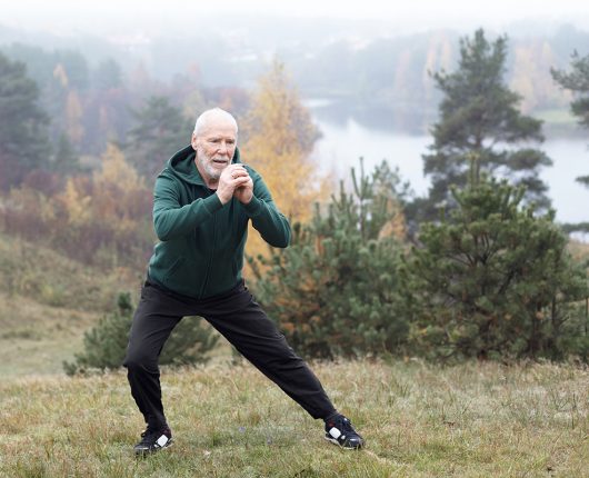 Persona de la tercera edad practica un estiramiento en el bosque, previo a iniciar su actividad física. 