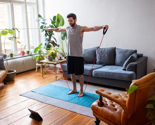 Hombre haciendo ejercicio con banda elástica en casa con una tableta digital. 